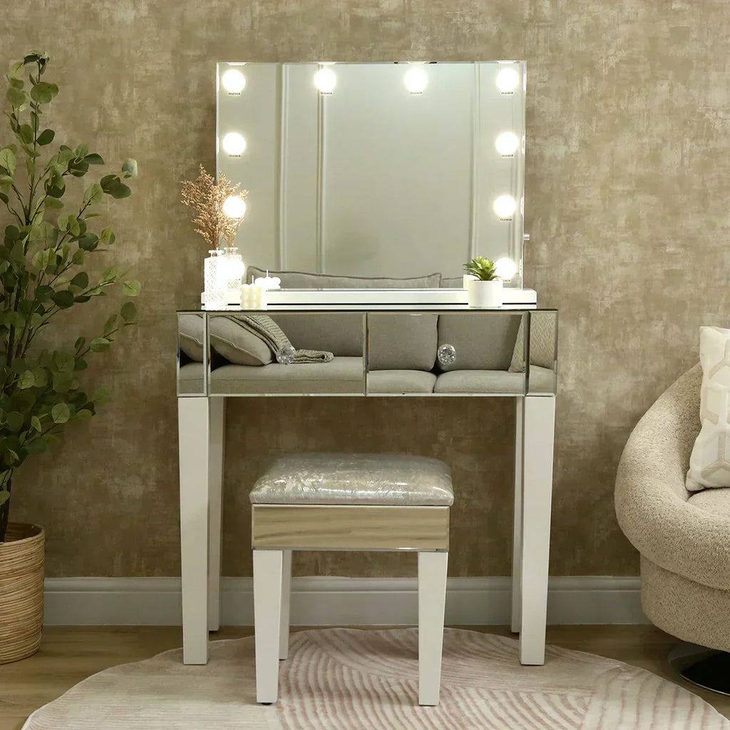 A chic mirrored dressing table with illuminated Hollywood-style bulbs, paired with a cushioned stool and styled with candles, decor pieces, and a small plant against a textured beige wall.