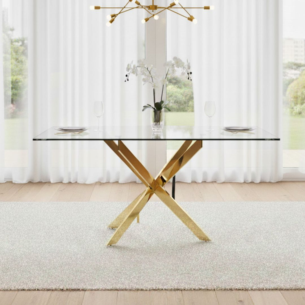 Dining table with gold legs and glass top in a room with large windows and white curtains.